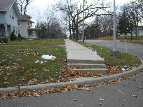 Steps and a non ADA compliant curb cut in Flint, Michigan make this sidewalk impossible to use by anyone requiring a mobility device.  Photo by Barbara McCann
